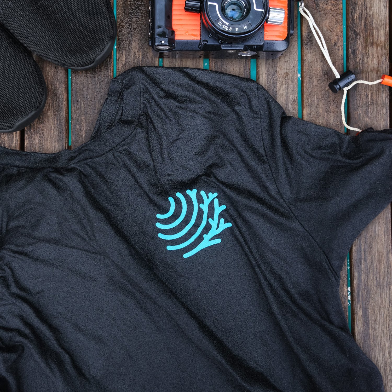 The FishEye Collaborative logo in teal is seen on a black tee-shirt, soaking wet and atop a deck above the water, surrounded by water shoes and a vintage Nikonos underwater camera