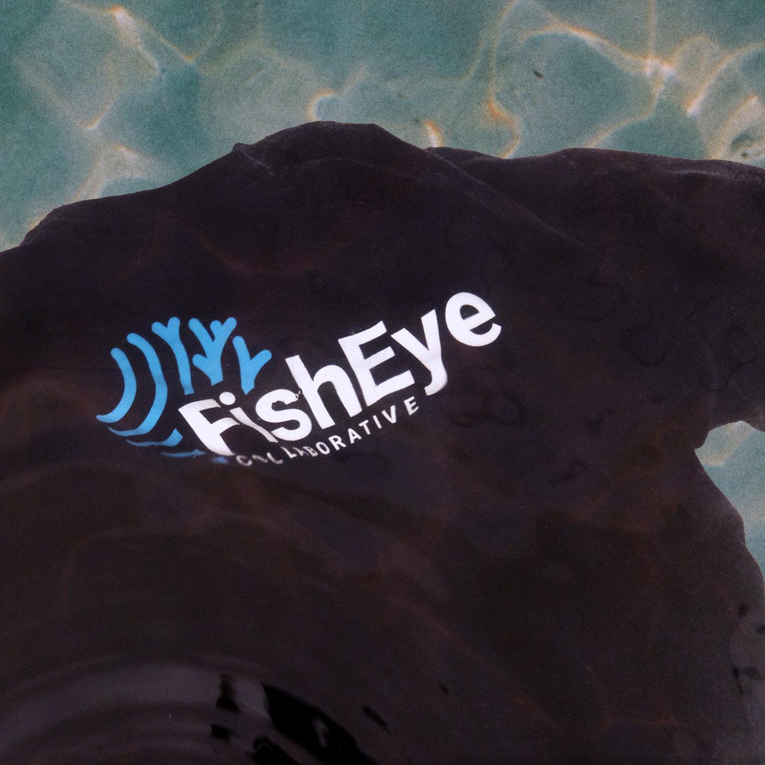 A black t-shirt with the FishEye Collaborative logo is seen gently floating submerged in the shallow tropical ocean