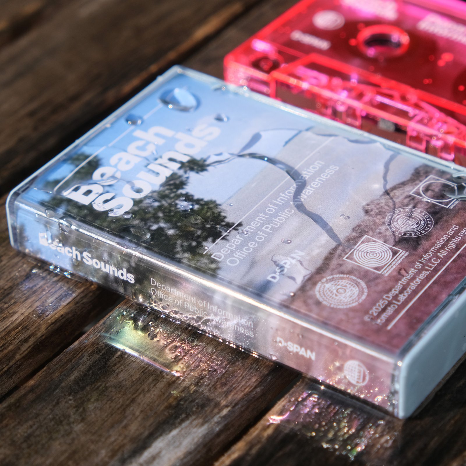 A close shot of Beach Sounds cassette tape case splashed with reflective water on a wood slat deck