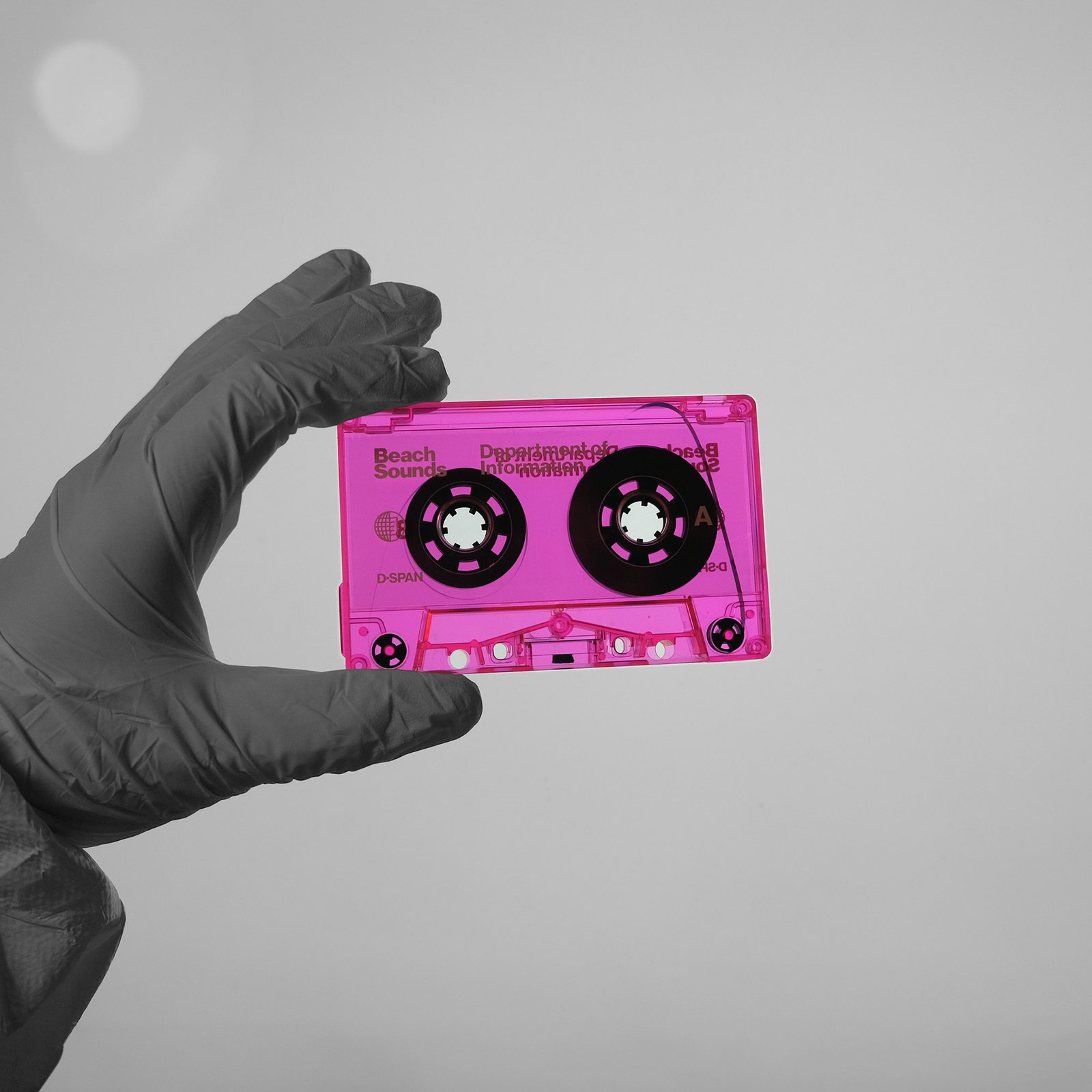 A white-suited hand in a laboratory holds up a copy of the fluorescent pink cassette tape Beach Sounds
